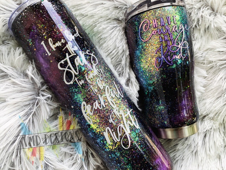 May include: Two stainless steel tumblers with a galaxy-inspired design. The larger tumbler has the text "I have loved the stars too fondly to be fearful of the night". The smaller tumbler has the text "Have courage and be kind".
