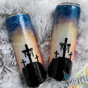 May include: Two stainless steel tumblers with a glitter finish. The tumblers feature a sunset gradient with a silhouette of three crosses on a hill. The tumblers are perfect for holding hot or cold beverages.