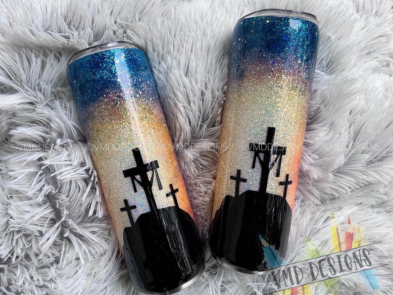 May include: Two stainless steel tumblers with a glitter finish. The tumblers feature a sunset gradient with a silhouette of three crosses on a hill. The tumblers are perfect for holding hot or cold beverages.