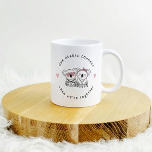 May include: White ceramic mug with a handle, featuring a design of two koalas with hearts and the text "OUR HEARTS connect when we're together". The mug is on a wooden round surface.