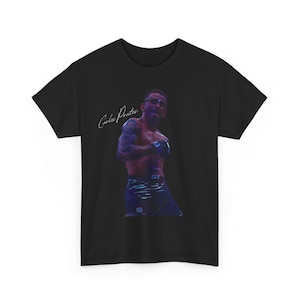 May include: Black t-shirt featuring a vibrant image of a person in a fighting stance, wearing gloves and shorts. The image includes the text "Carlos Prates" in a cursive font. The UFC logo is visible on the shorts.