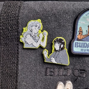 Może przedstawiać: Dwie emaliowane przypinki w stylu anime przedstawiające kobiety w pozach walki. Przypinki mają żółtą obwódkę i są głównie szare, czarne i białe. Jedna przypinka przedstawia kobietę z długimi włosami, druga z krótkimi.