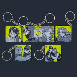 Op de afbeelding: Sleutelhangers met illustraties van mensen in verschillende poses, elk bevestigd aan een zilveren sleutelring. De sleutelhangers hebben een neongele achtergrond. De illustraties zijn in zwart en wit, met een hartontwerp op een sleutelhanger.