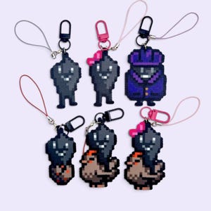Op de afbeelding: Zes pixel art sleutelhangers met donkergrijze roetwezentjes. Sommige hebben roze strikjes, één draagt een paarse jas en andere zitten op kleine bruine wezentjes. Elke sleutelhanger is voorzien van een koordje en een plastic sluiting.