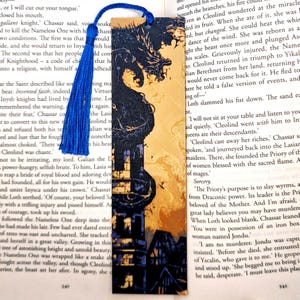 May include: A bookmark featuring a black dragon with a yellow background. The dragon is flying up towards a dark, gothic-style building. The bookmark is made of wood and has a blue tassel.