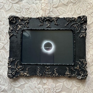 May include: Black ornate picture frame with a gold-colored design. The frame holds a black and white photograph of a solar eclipse.