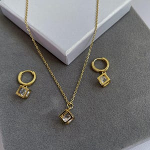 May include: A gold necklace and earrings set with a square pendant and matching earrings. Each piece features a clear cubic zirconia stone in a gold frame.