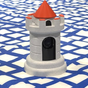 May include: A gray toy castle tower with a red conical roof. The tower has a cylindrical body with a rectangular opening and small rectangular crenellations. The base is a wider, circular shape. The background is a blue and white patterned surface.
