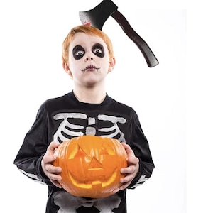Puede incluir: Un niño vestido de esqueleto para Halloween sostiene una calabaza tallada. Un hacha de juguete con una mancha roja está en la cabeza del niño.