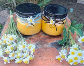 May include: Two yellow candles in glass jars with black lids. The jars are decorated with blue and white ribbon, a bee, and a honeycomb design. The candles are sitting on a wooden surface with a bouquet of white daisies.
