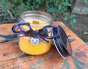 May include: A yellow candle in a glass jar with a black lid. The jar is decorated with a purple ribbon and a black and white cat charm. The candle is sitting on a wooden surface.