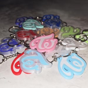 Puede incluir: Una colección de llaveros coloridos con la palabra "Twice" en una fuente estilizada. Los llaveros son de varios colores, incluyendo rosa, azul, rojo y verde, con llaveros plateados.