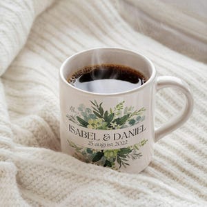K&ouml;nnte beinhalten: Wei&szlig;e Keramiktasse mit gr&uuml;nem Blumendesign und dem Text "Isabel & Daniel 25. August 2022". Die Tasse ist mit Kaffee gef&uuml;llt und Dampf steigt aus der Oberseite auf.