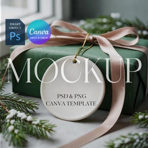 Puede incluir: Maqueta de un adorno redondo de cerámica blanca con la palabra "MOCKUP" en letras grandes. El adorno está frente a una caja de regalo verde envuelta con una cinta de satén rosa claro. El texto "PSD & PNG CANVA TEMPLATE" está debajo. Follaje verde en el fondo.
