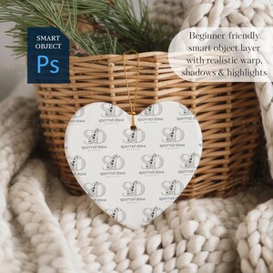 May include: Heart-shaped ceramic ornament with a repeating pattern of a logo and the text "SPOTTED DANI". The ornament hangs from a gold string. The background includes a woven basket, greenery, and a soft, white blanket.