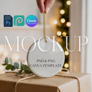 Puede incluir: Adorno blanco y redondo con una cuerda dorada, sostenido por una mano. La palabra "MOCKUP" en letras blancas grandes. Debajo, las palabras "PSD & PNG CANVA TEMPLATE". El adorno está sobre un regalo envuelto.