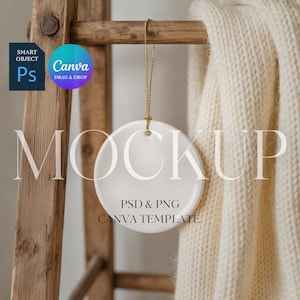 Op de afbeelding: Een witte, ronde keramische ornament hangt aan een gouden draad tegen een houten ladder en een crèmekleurige deken. Het ornament heeft de tekst "MOCKUP", evenals "PSD & PNG" en "CANVA TEMPLATE".