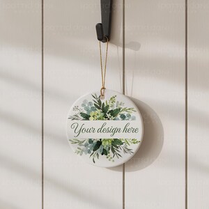 May include: A circular, white ceramic ornament featuring a floral design in green and blue hues. The ornament has the text "Your design here" across the middle. It is suspended by a gold string and a black hook.