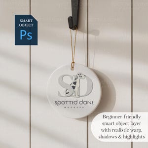 May include: A round white ceramic ornament featuring a Dalmatian dog design and the text "Spotted Dane Mockups." The ornament is suspended by a gold string. The image also includes a "Smart Object Ps" icon and text: "Beginner-friendly smart object layer with realistic warp, shadows & highlights."