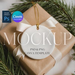 May include: A white ceramic ornament with a gold string hangs over a wrapped gift. The gift is wrapped in brown paper and tied with a cream-colored ribbon. The word "MOCKUP" is printed on the ornament. The image also includes text that says "PSD & PNG CANVA TEMPLATE".