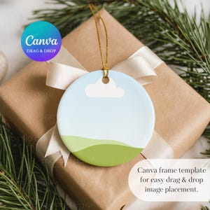 May include: A round ceramic ornament with a sky and landscape design. The ornament has a blue sky, a white cloud, and green hills. The ornament is hanging by a gold string. The text "Canva" and "Drag & Drop" are visible.