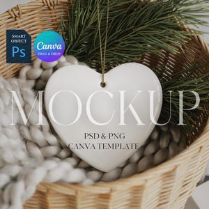 May include: A white heart-shaped ornament with a gold string, presented in a wicker basket with a soft, grey blanket and pine branches. The ornament has the word "MOCKUP" printed on it, along with "PSD & PNG CANVA TEMPLATE".