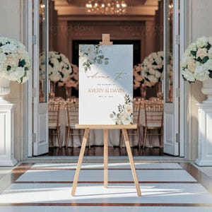 May include: A white wedding welcome sign with gold lettering and floral accents. The sign reads "Welcome to the wedding of Avery & Davis, May 12, 2024". The sign is displayed on a wooden easel.