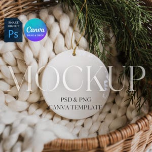 May include: A white ceramic round ornament with a gold string, featuring the word "MOCKUP" and the text "PSD & PNG CANVA TEMPLATE". The ornament is set in a woven basket with a white knitted blanket and evergreen branches.