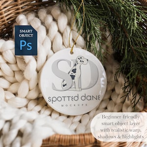 May include: A white ceramic ornament with a Dalmatian dog design and the text "Spotted Dane Mockups." The ornament hangs from a gold string. The background includes a white knitted blanket and greenery.