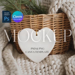May include: A white heart-shaped ornament with a gold string for hanging. The ornament is in front of a woven basket and a cozy, off-white knitted blanket. The text "MOCKUP" is displayed on the ornament, along with "PSD & PNG CANVA TEMPLATE".