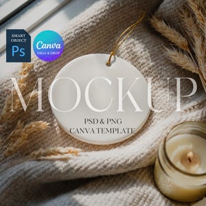 Puede incluir: Un adorno redondo de cerámica blanca con una cuerda dorada, con la palabra "MOCKUP" en una fuente suave. También se ve el texto "PSD & PNG CANVA TEMPLATE". El adorno descansa sobre una superficie beige texturizada, con una vela encendida y espigas de trigo.