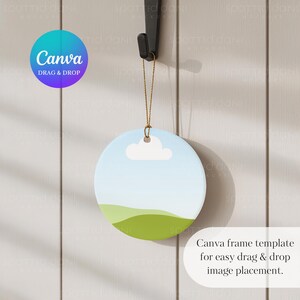 May include: A circular ceramic ornament with a sky and landscape design, suspended from a black hook. The ornament depicts a blue sky, a white cloud, and green hills. Text on the image says "Canva DRAG & DROP" and "Canva frame template for easy drag & drop image placement."