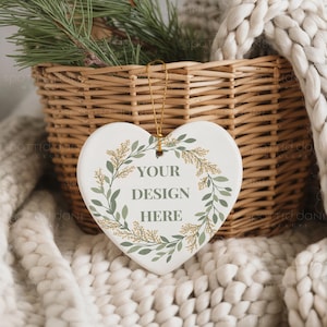 May include: A heart-shaped ceramic ornament with the text "YOUR DESIGN HERE" surrounded by a green and gold botanical wreath. The ornament hangs from a gold string, set against a woven basket and a cozy, off-white blanket.