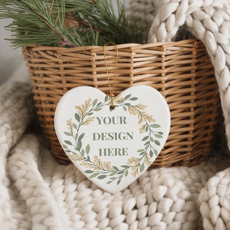 May include: A heart-shaped ceramic ornament with the text "YOUR DESIGN HERE" surrounded by a green and gold botanical wreath. The ornament hangs from a gold string, set against a woven basket and a cozy, off-white blanket.