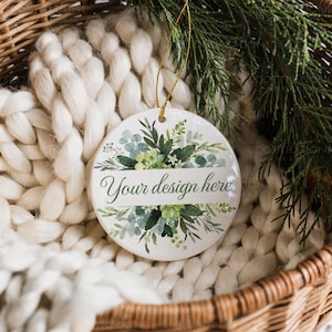 May include: A white ceramic ornament with a gold string, featuring a floral design in shades of green and blue. The text "Your design here" is displayed on a white banner. The ornament is set against a backdrop of a woven basket and a white knitted blanket.