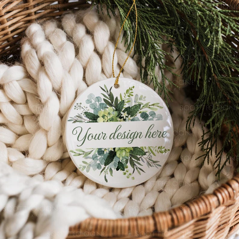 May include: A white ceramic ornament with a gold string, featuring a floral design in shades of green and blue. The text "Your design here" is displayed on a white banner. The ornament is set against a backdrop of a woven basket and a white knitted blanket.
