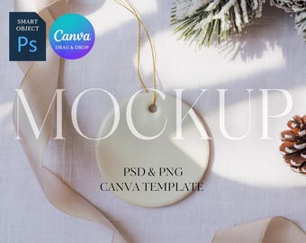 Puede incluir: Un adorno de cerámica redondo, color crema, con una cuerda dorada, ideal para la decoración navideña. La palabra "MOCKUP" se muestra en letras grandes y elegantes. También se ve el texto "PSD & PNG CANVA TEMPLATE". El fondo incluye piñas y una cinta.