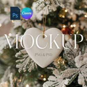 May include: White ceramic heart ornament hanging on a Christmas tree with snow-covered branches and twinkling lights. The ornament has the text "MOCKUP PNG & PSD" printed on it.