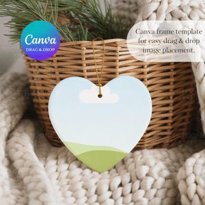 May include: Heart-shaped ornament with a sky and landscape design. The ornament has a light blue sky, a white cloud, and green hills. It hangs from a gold string. The text "Canva DRAG & DROP" is visible.