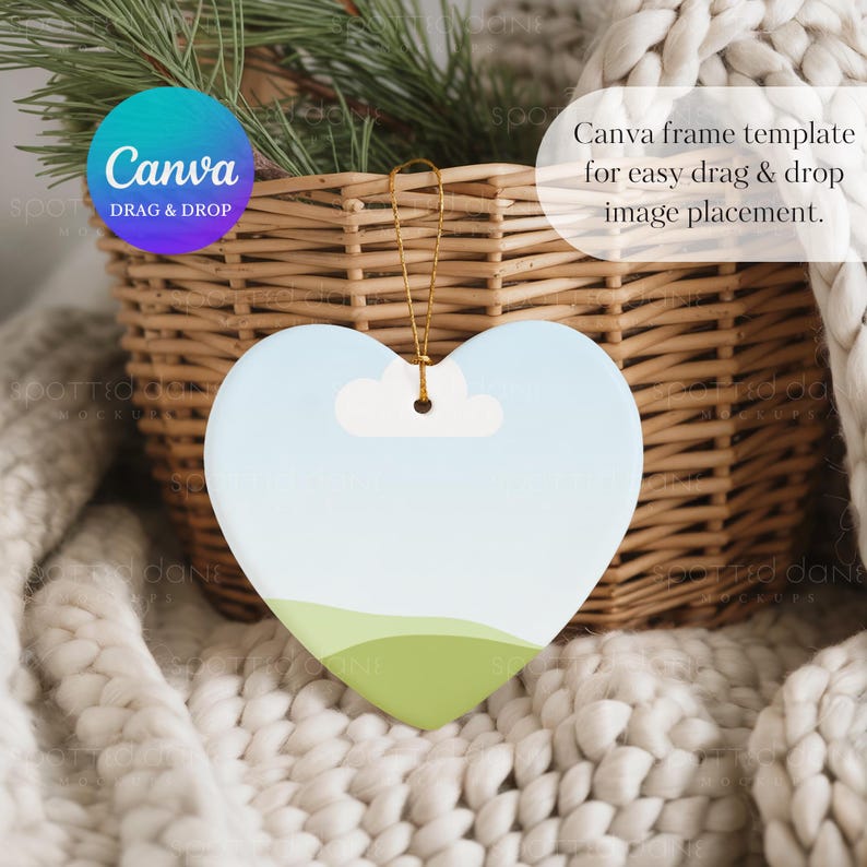 May include: Heart-shaped ornament with a sky and landscape design. The ornament has a light blue sky, a white cloud, and green hills. It hangs from a gold string. The text "Canva DRAG & DROP" is visible.
