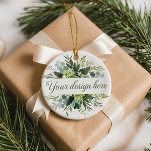 May include: A white ceramic ornament with a gold string, featuring a floral design in shades of green and blue. The ornament has the text "Your design here" in a banner. It rests on a wrapped gift box with a cream ribbon, surrounded by pine branches.