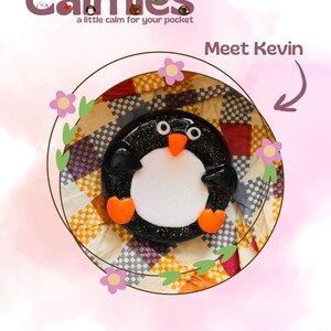 Può includere: Un oggetto rotondo a forma di pinguino bianco e nero con ali nere glitterate, pancia bianca, piedi arancioni e becco arancione. Il testo Calmies a little calm for your pocket e Meet Kevin è visibile. Incorniciato da un sottile cerchio marrone.