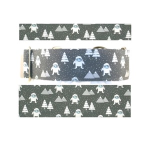 May include: A gray dog collar featuring a repeating pattern of white cartoon yetis, pine trees, and mountains. The collar has a metal buckle and a D-ring for leash attachment. The background is a dark gray with white dots.
