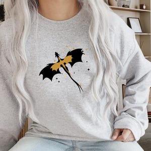 May include: A light grey sweatshirt with a gold and black dragon design and star accents. The dragon has outstretched wings and a long tail. The sweatshirt is a crew neck style and has long sleeves.