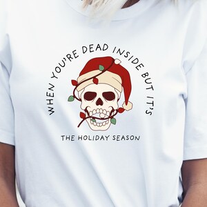 May include: White t-shirt with a graphic of a skull wearing a Santa hat and Christmas lights. The text around the skull reads "When you're dead inside but it's the holiday season."