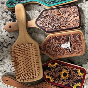 Puede incluir: Una colección de cinco cepillos para el cabello rectangulares con mangos de madera. Cuatro cepillos presentan diseños de cuero repujado con motivos florales o de cráneo de toro, incluyendo tonos turquesa, bronceado natural y girasoles. Un cepillo muestra las cerdas.