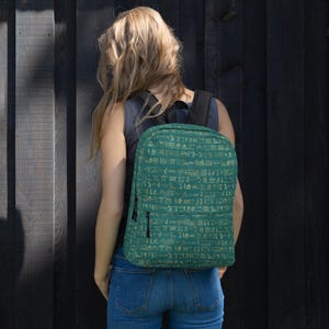 May include: Teal backpack with a repeating pattern of gold hieroglyphs. The backpack has a black zipper and straps. The person in the image is wearing blue jeans and a gray top.