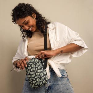 May include: A black crossbody bag with a green and white money print. The bag has a long adjustable strap and a zipper closure.