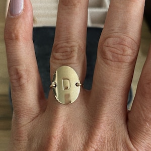 May include: A gold ring with a round, oval-shaped face featuring the letter "D" in a raised design.