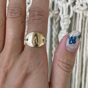 May include: A gold ring with a hammered oval-shaped band featuring the letter "A" in the center.
